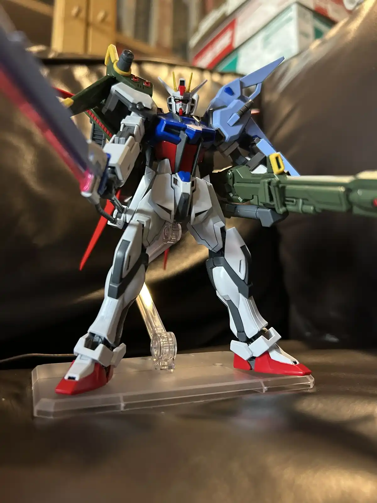 A detailed plastic model of a Gundam robot, primarily white with blue, red, and green accents, stands on a clear plastic…