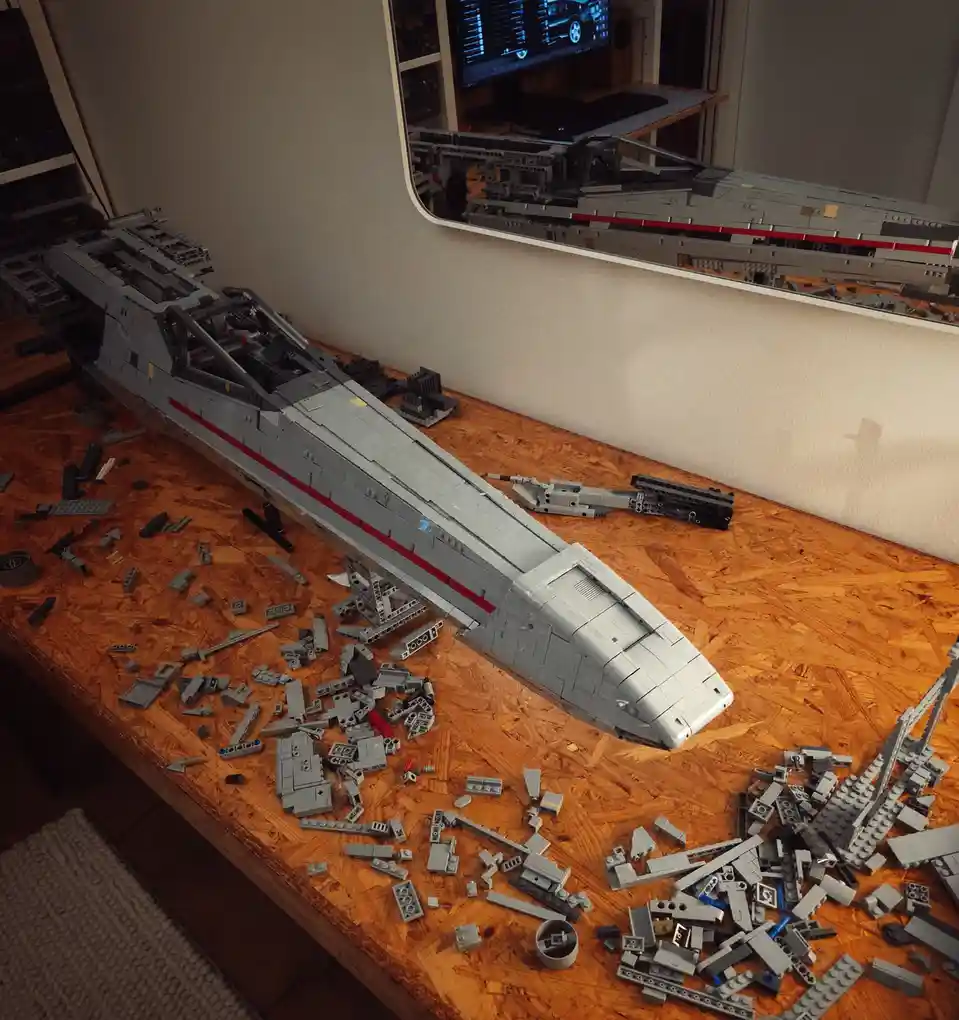 A partially assembled large grey Lego Star Wars spaceship sits on a wooden table, surrounded by scattered Lego pieces. A…
