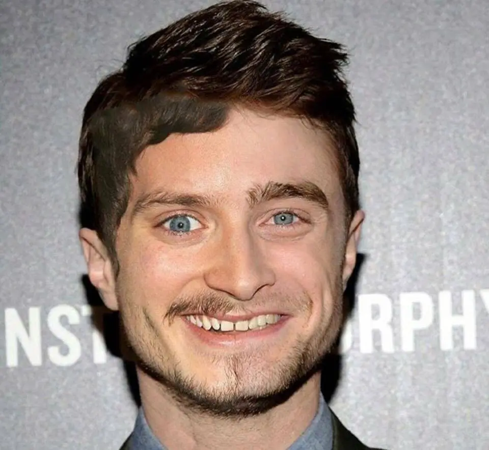 A close-up photo of actor Daniel Radcliffe smiling broadly with his teeth showing, against a gray backdrop with partial…