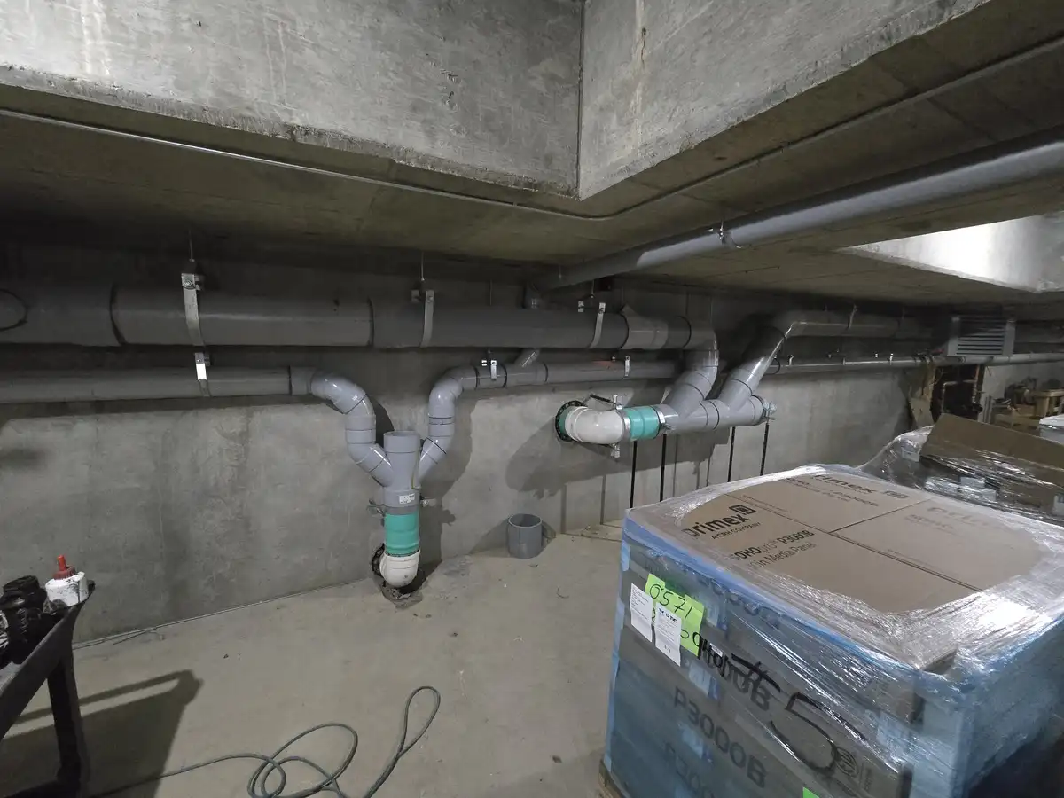A photo of a concrete basement with grey PVC pipes running along the walls and ceiling, some with green fittings. A large…