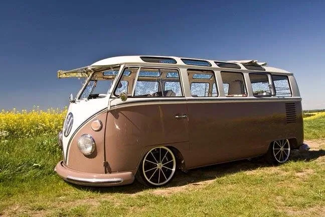 A vintage brown and white Volkswagen Type 2 van with custom chrome wheels is parked in a grassy field next to a yellow…