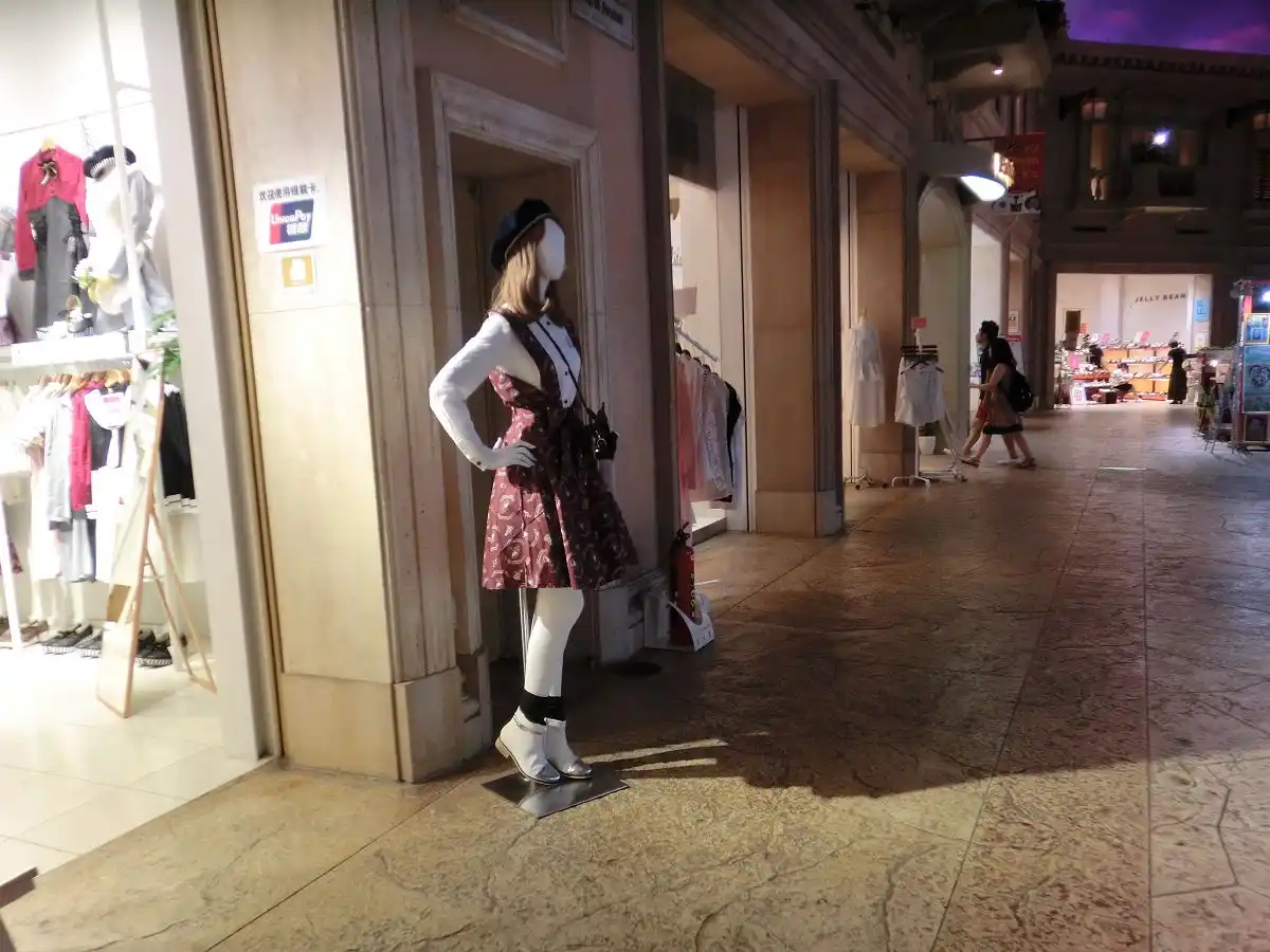 A mannequin in a shopping mall corridor is dressed in a patterned dress, white tights, and boots, wearing a black beret and…