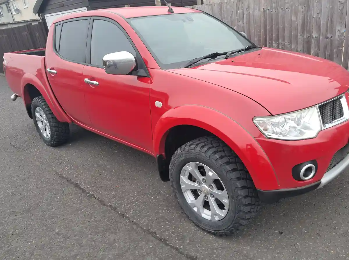 A red Mitsubishi L200 pickup truck with large off-road tires is parked on asphalt in front of a wooden fence. The overlaid…