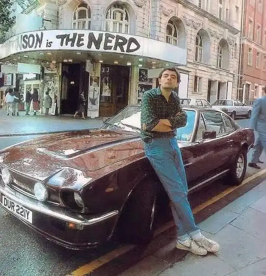 A man in a plaid shirt and jeans leans against a dark brown vintage car parked on a city street. Behind him, a theater…