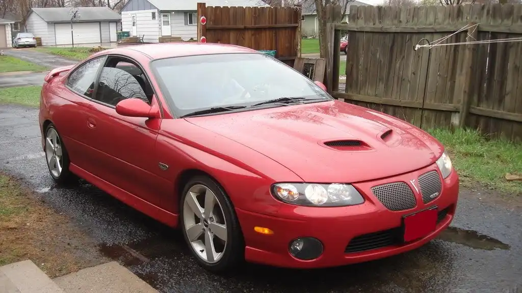 a red 2004 pontiac grand am gt is parked on a wet driveway in front of a wooden fence, with a residential neighborhood in…