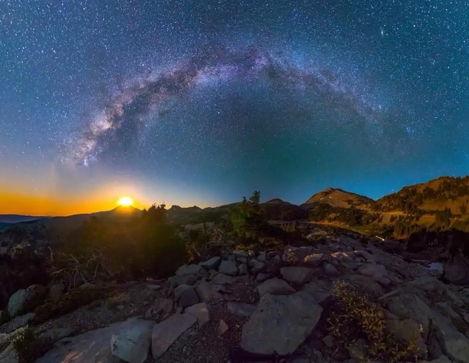 A stunning wide-angle night photograph captures the Milky Way galaxy arching across a star-filled sky, with a bright sunrise…