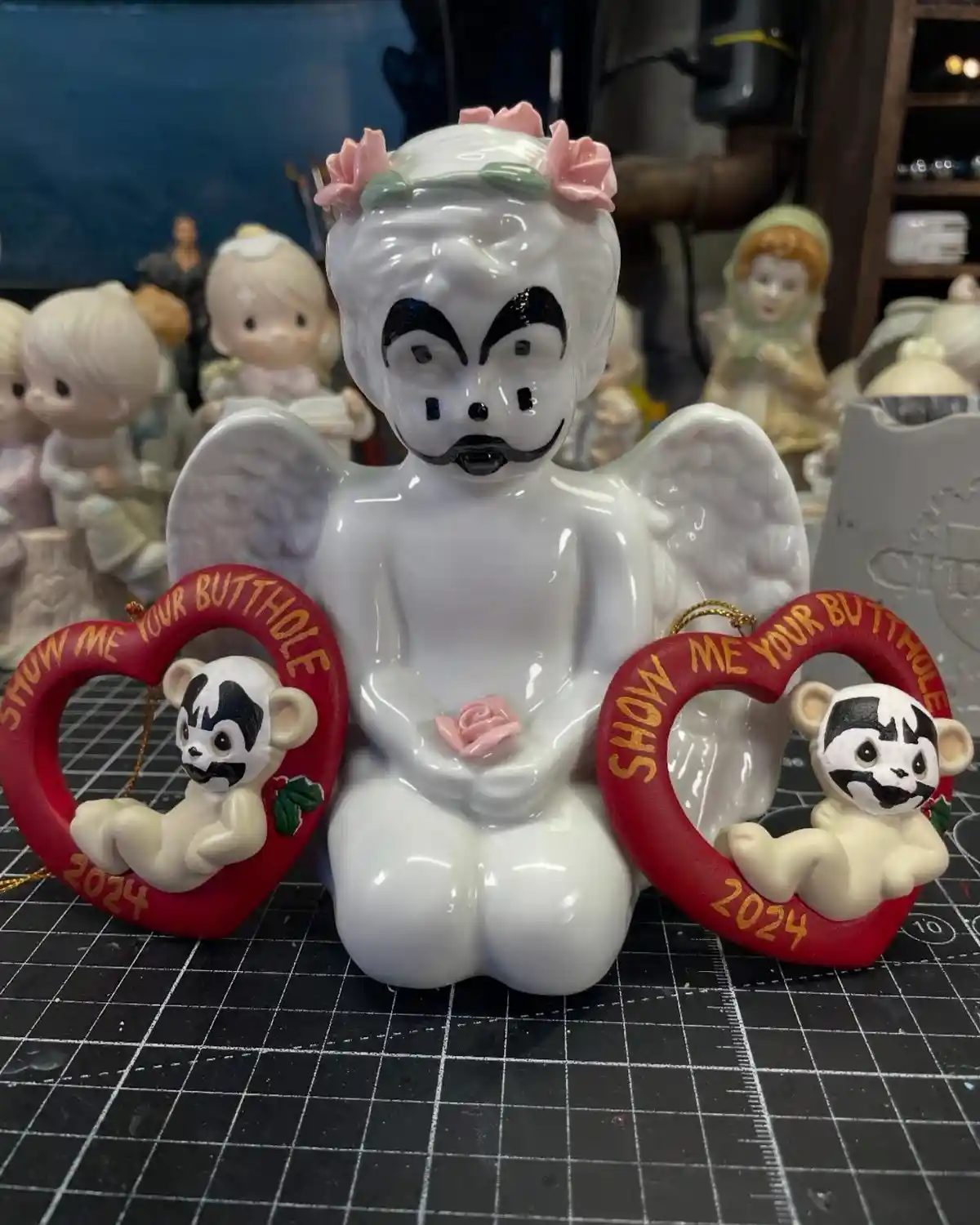 A white ceramic angel figurine with a black mustache and angry eyebrows sits between two red heart-shaped ornaments. Each…
