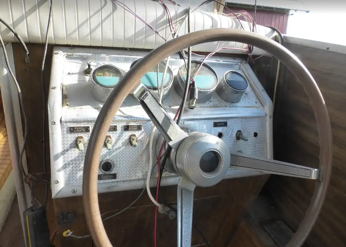 A close-up photo of a vintage boat's control panel with a wooden steering wheel, multiple gauges, and exposed wiring. The…