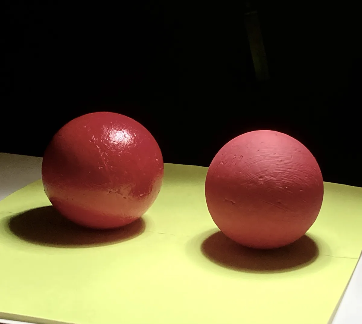 Two red spheres sit side-by-side on a bright yellow surface against a black background. The sphere on the left is a glossy…