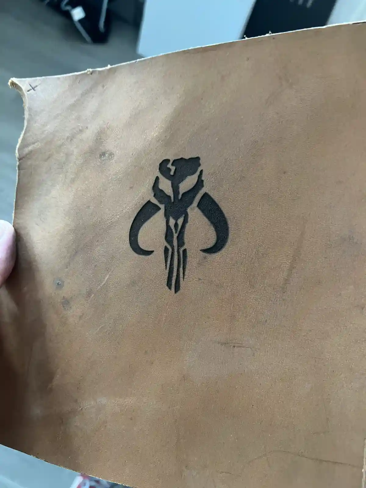 A close-up photo shows a hand holding a piece of brown, textured leather with a black inked Mandalorian symbol printed on…