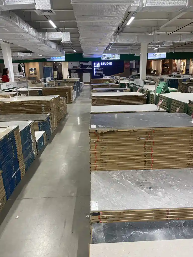 An aisle in a large tile store filled with stacks of large, flat, rectangular tiles in various shades of gray and white…