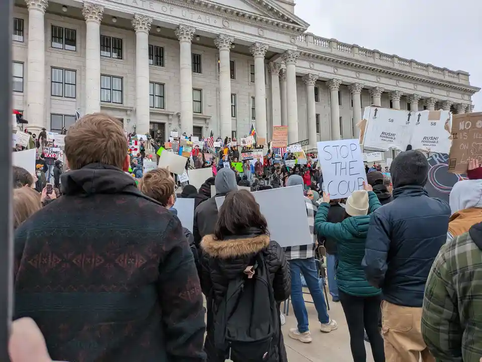 A crowd of protesters stands in front of the Utah State Capitol building, holding various handmade signs. One prominent sign…