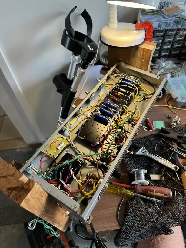 A cluttered workbench shows an open electronic device with colorful wires, resistors, and capacitors. Tools like a wrench…