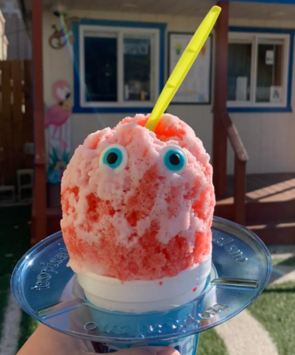 A pink and white snow cone in a white cup sits on a blue plastic tray, topped with two large googly eyes and a yellow straw…