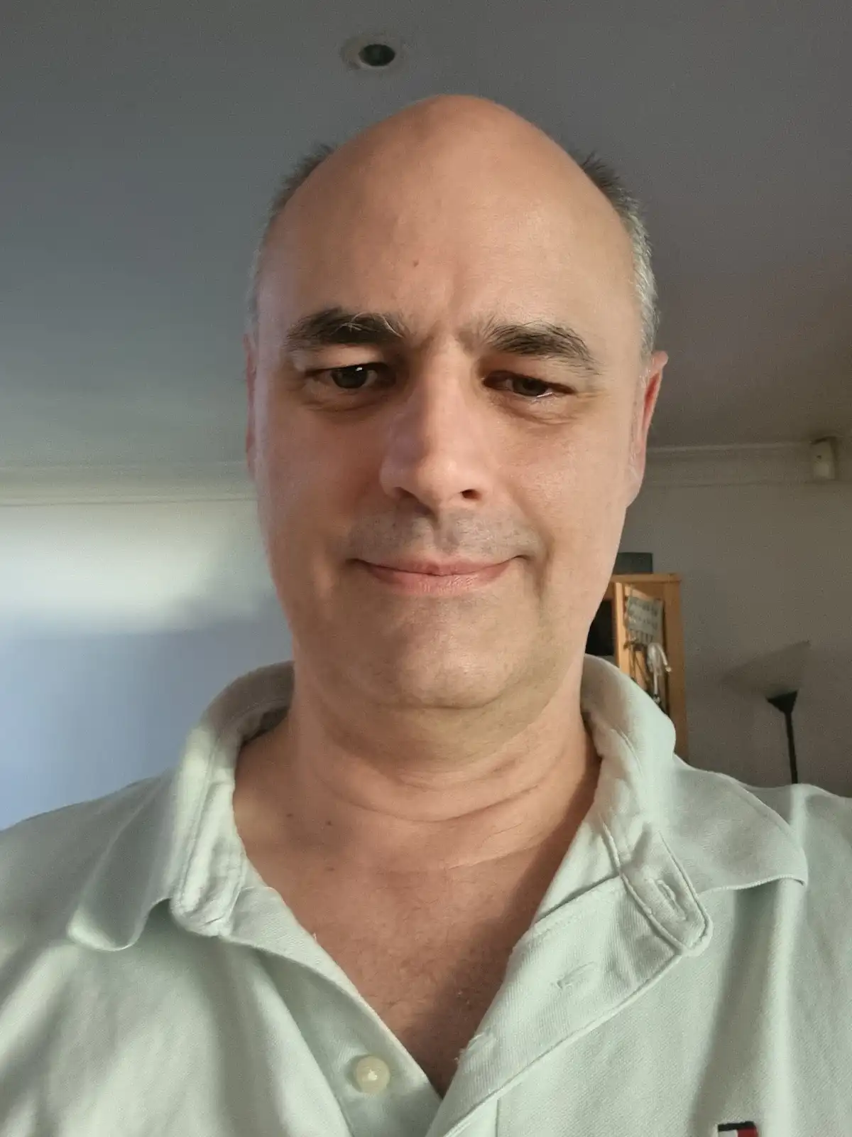 A close-up selfie of a middle-aged man with a balding head and gray hair on the sides, wearing a light green polo shirt with…
