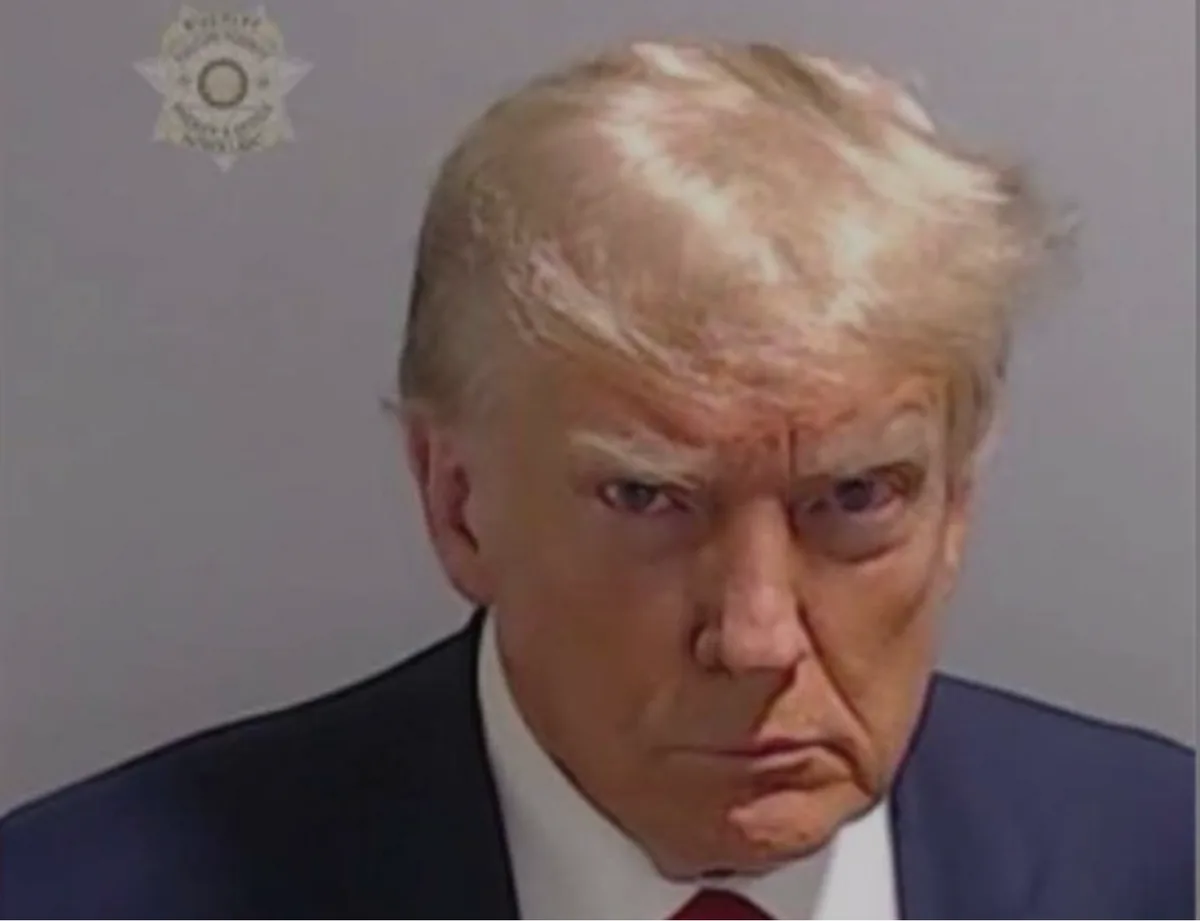 A mugshot-style photo of Donald Trump with a stern expression, wearing a dark suit and red tie, against a plain gray…