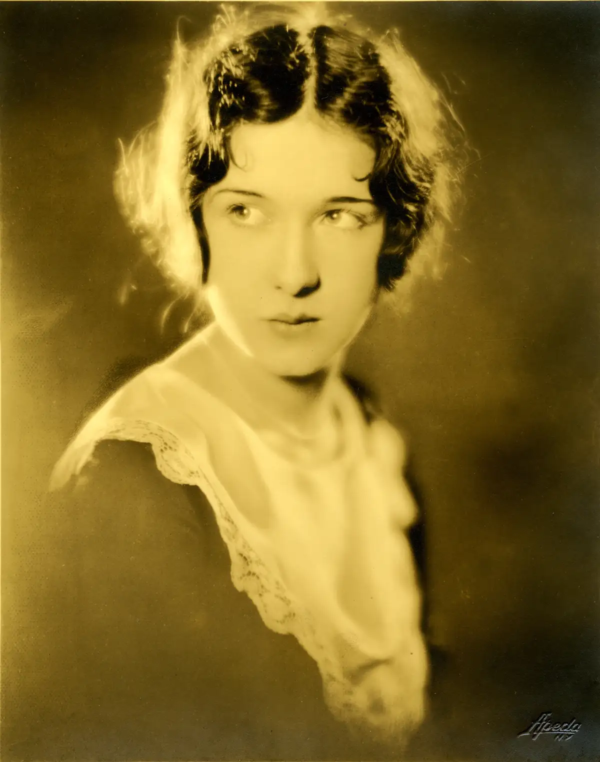 A sepia-toned vintage portrait of a young woman with short, wavy hair and a lace-trimmed collar, looking slightly away from…