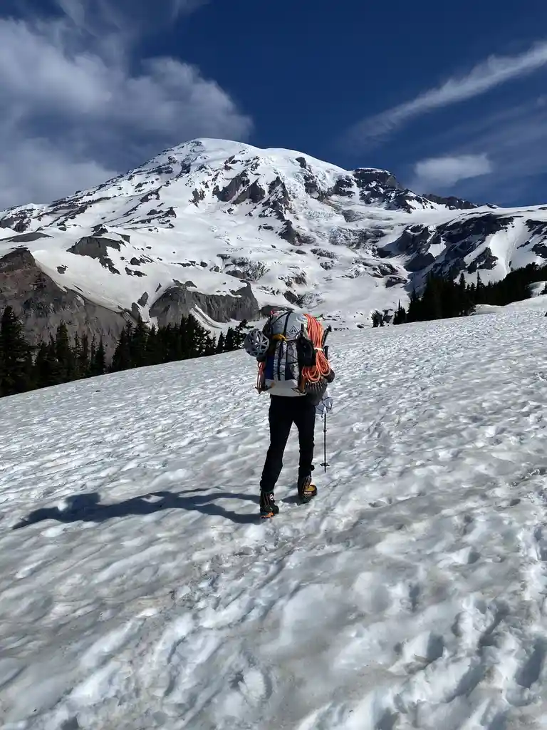 A person wearing a large backpack with climbing gear, including an orange rope, walks away from the camera across a snowy…