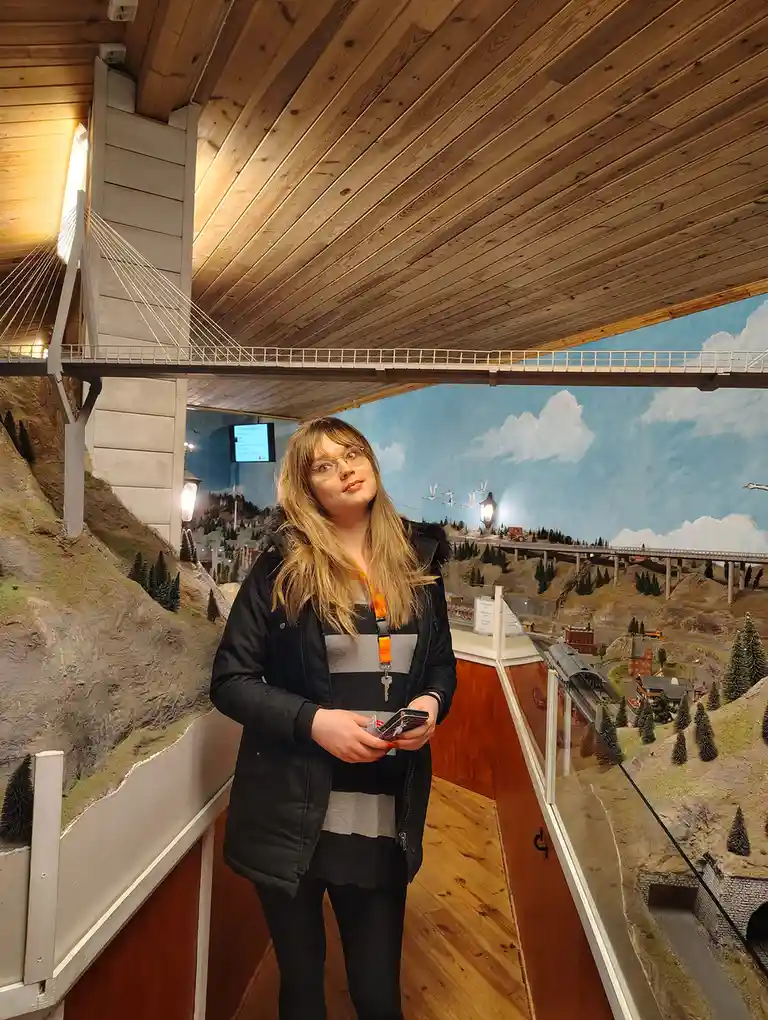 A young woman with glasses stands in a model train exhibit, looking upward with a mix of awe and apprehension. Behind her is…