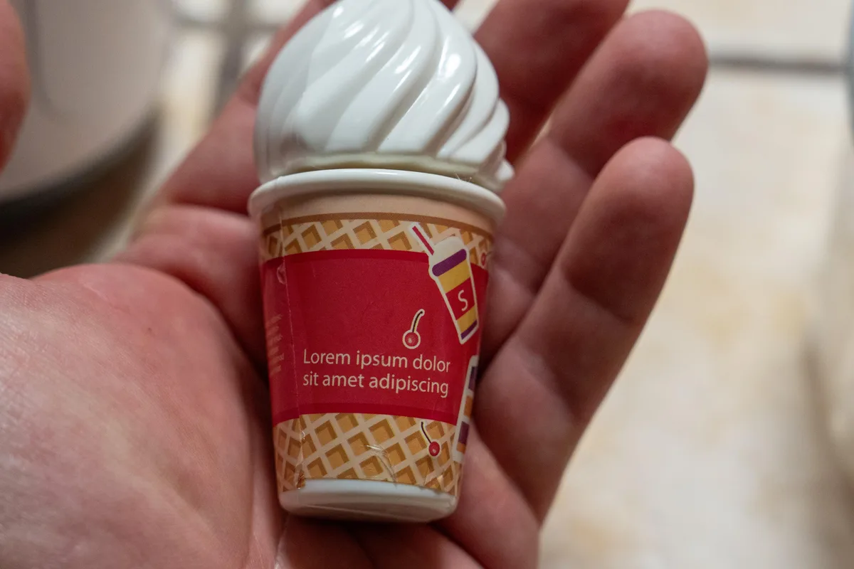 A hand holds a tiny, white, swirled soft-serve ice cream cup with a waffle-patterned sleeve and placeholder text. The cup is…