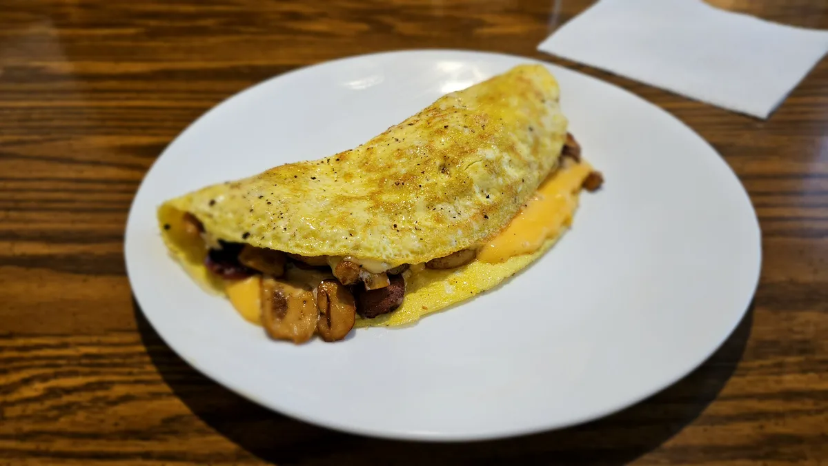 A close-up photo of a folded omelette on a white plate, filled with melted cheese and sautéed mushrooms and possibly…