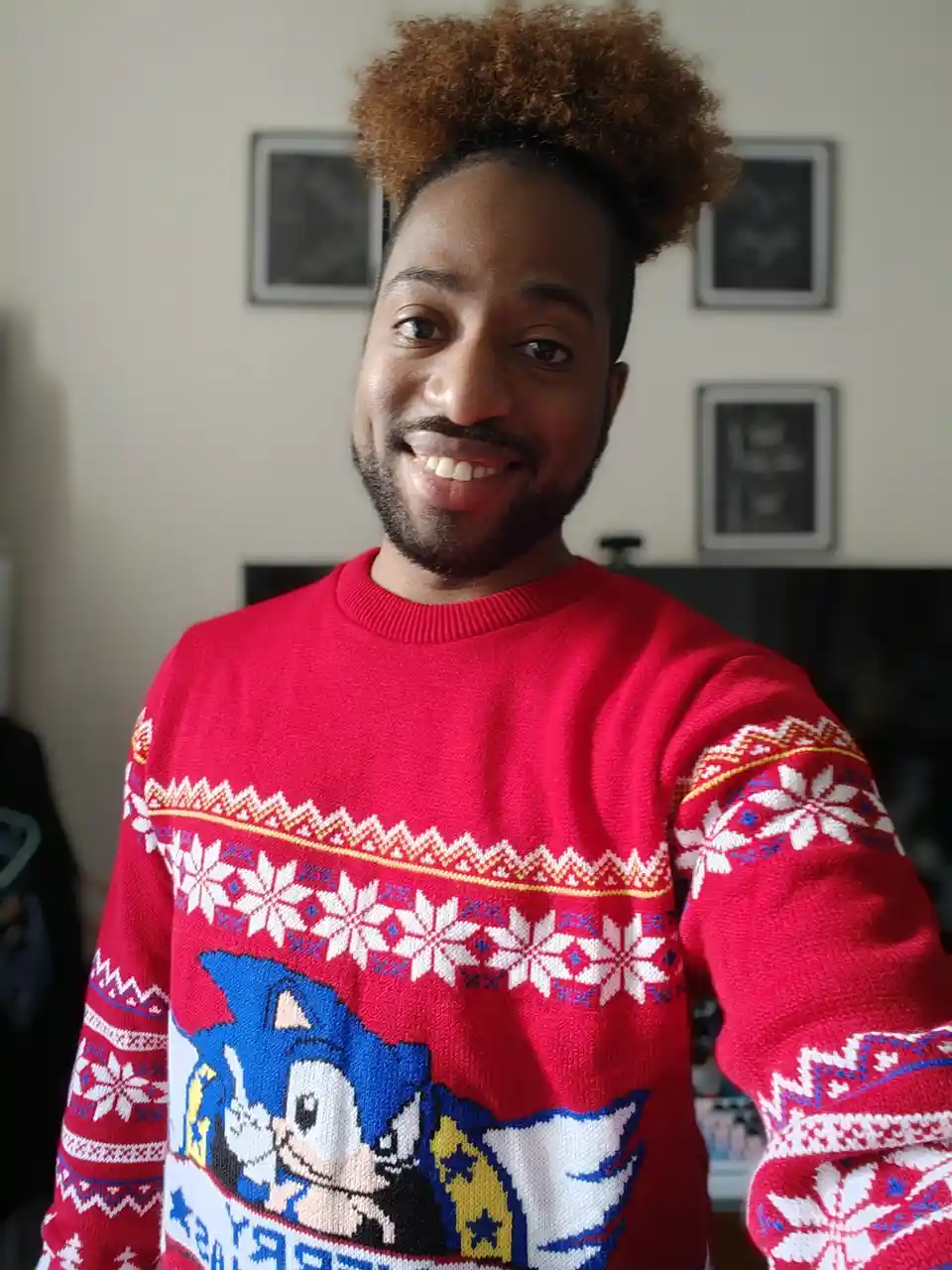A smiling person with curly hair wears a red, festive sweater featuring a large graphic of Sonic the Hedgehog surrounded by…
