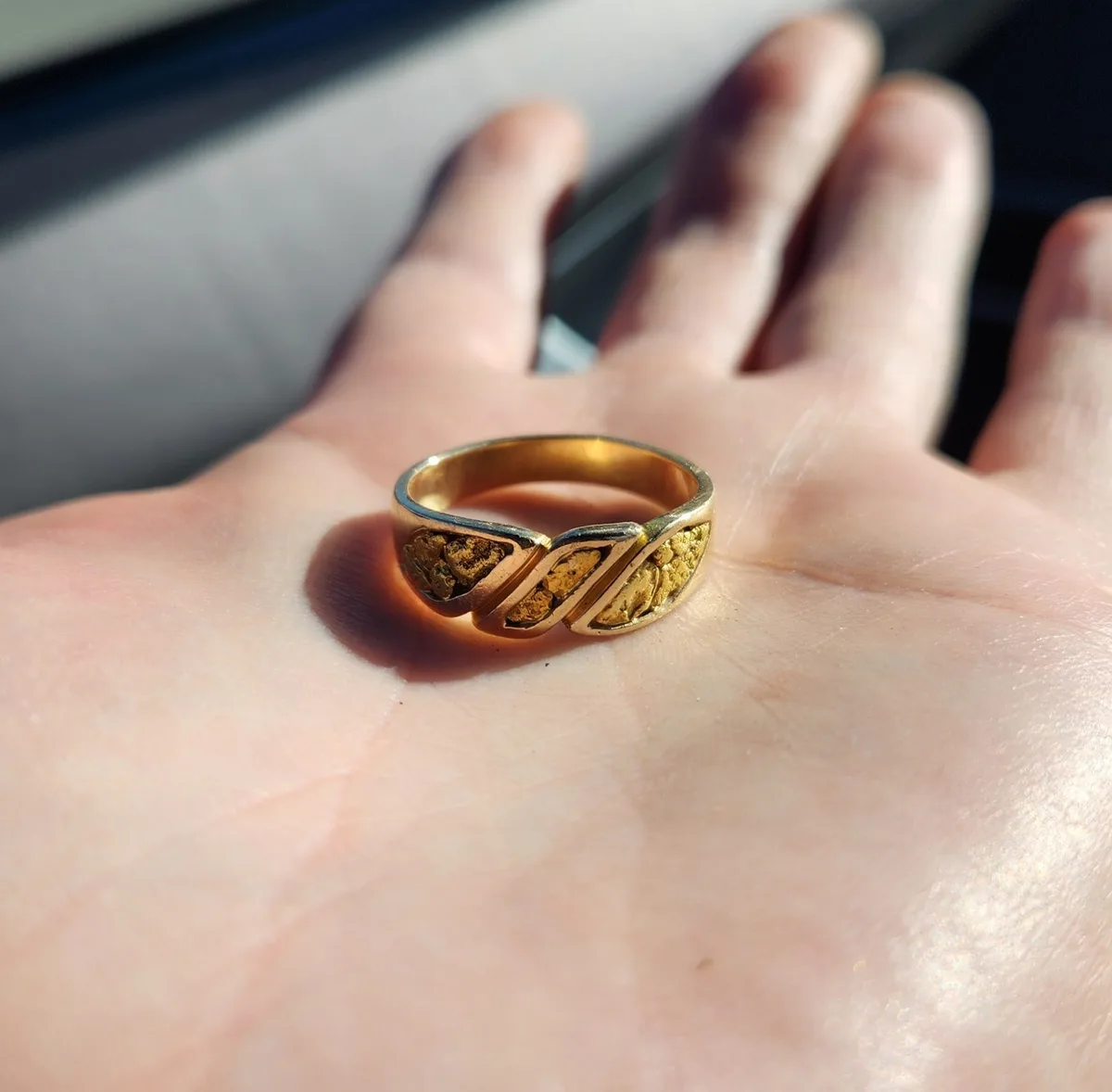 A close-up photo shows a gold ring with embedded gold nuggets resting on an open palm. The ring has a distinctive design…