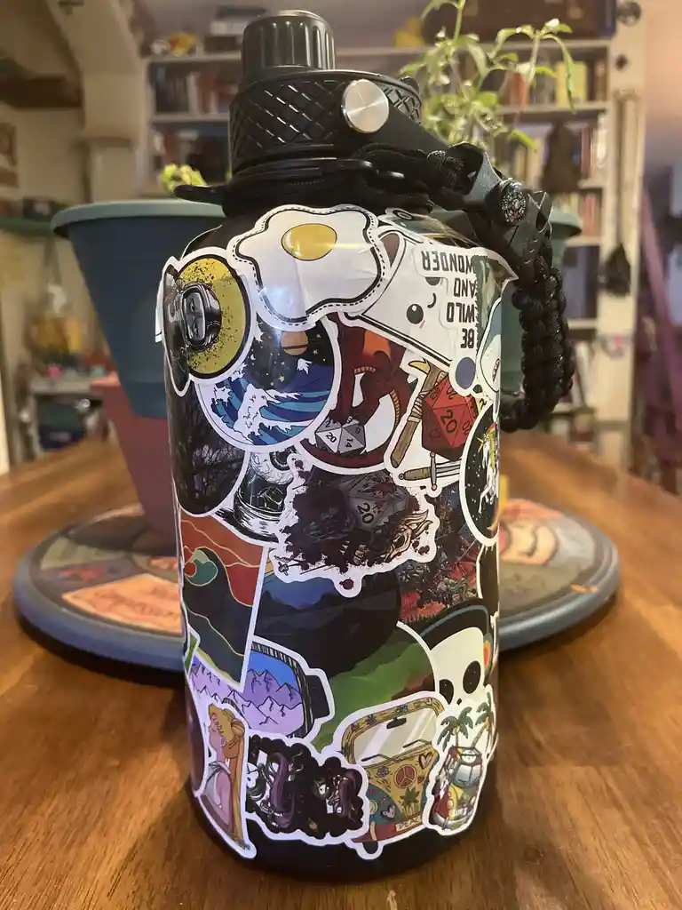 A black water bottle is covered in a dense collage of colorful stickers featuring diverse imagery including a fried egg, a…
