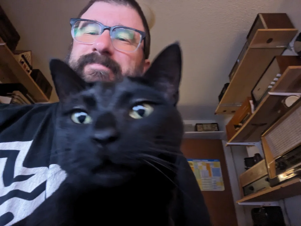 A man with glasses and a beard holds a black cat close to the camera, obscuring most of his face. The cat has wide…