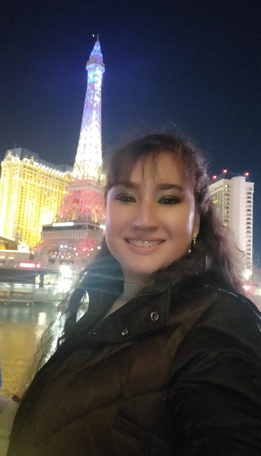 A smiling woman takes a selfie at night in front of the brightly lit replica of the Eiffel Tower at the Paris Las Vegas…