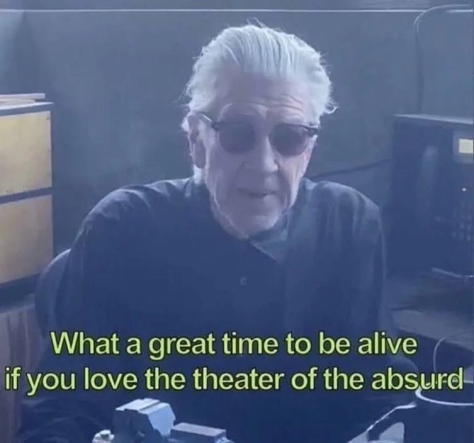 An older man with white hair and dark sunglasses sits in a dimly lit room with office equipment visible behind him. He has a…