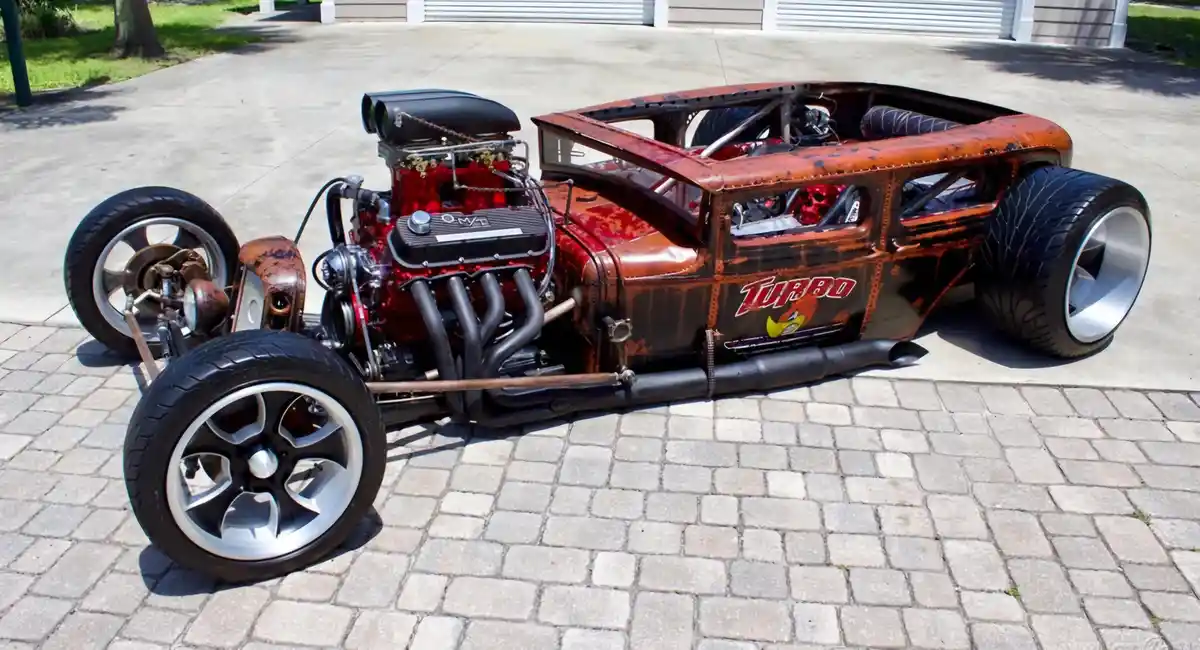 A custom-built hot rod with a red and black engine, exposed frame, and large tires sits on a paved driveway. The side of the…