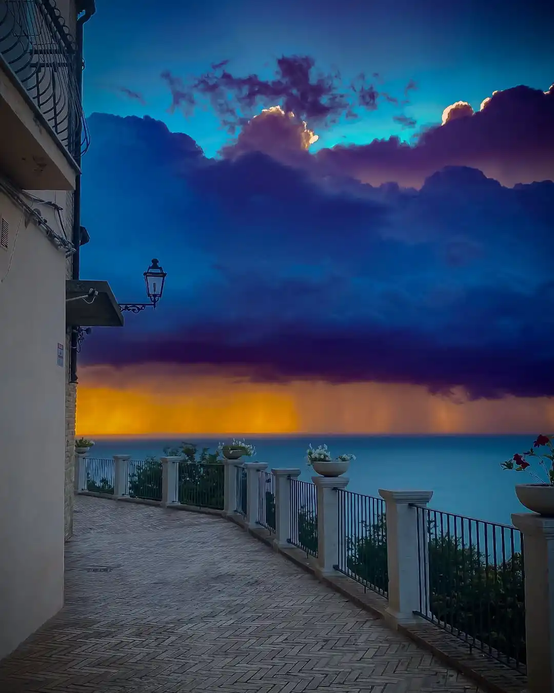 A scenic photo of a brick walkway overlooking the ocean at dusk, with dramatic purple and orange clouds above. A building…