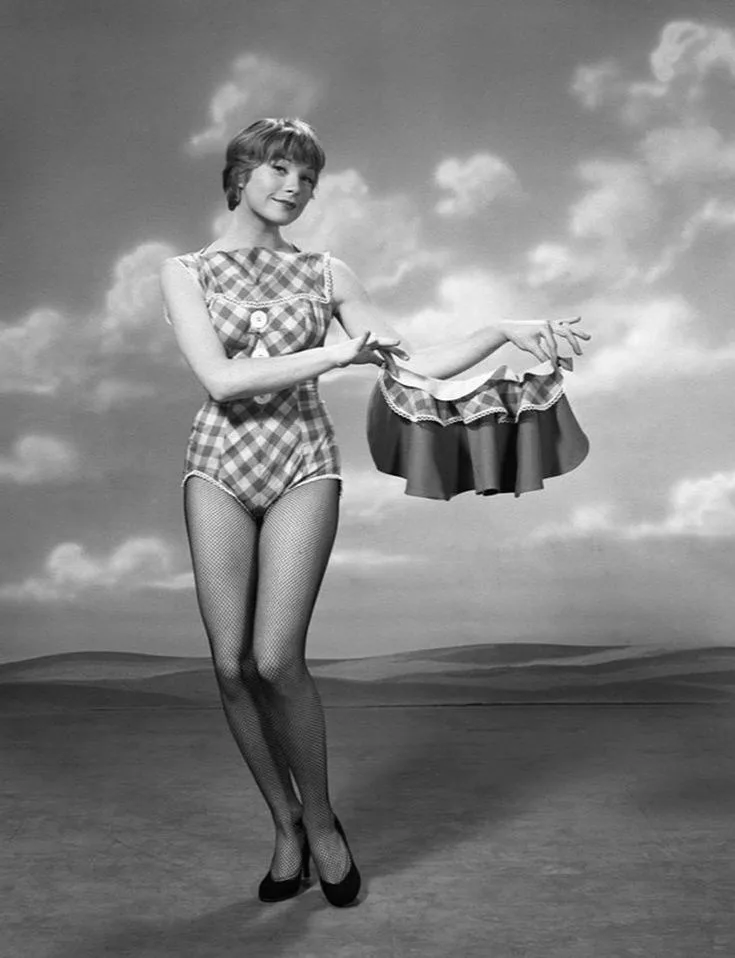 A black-and-white vintage photo shows a woman in a checkered one-piece swimsuit and fishnet stockings holding a detached…