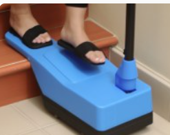 A person wearing black sandals stands on a blue footrest placed on a staircase, with a mop handle resting against the wall…