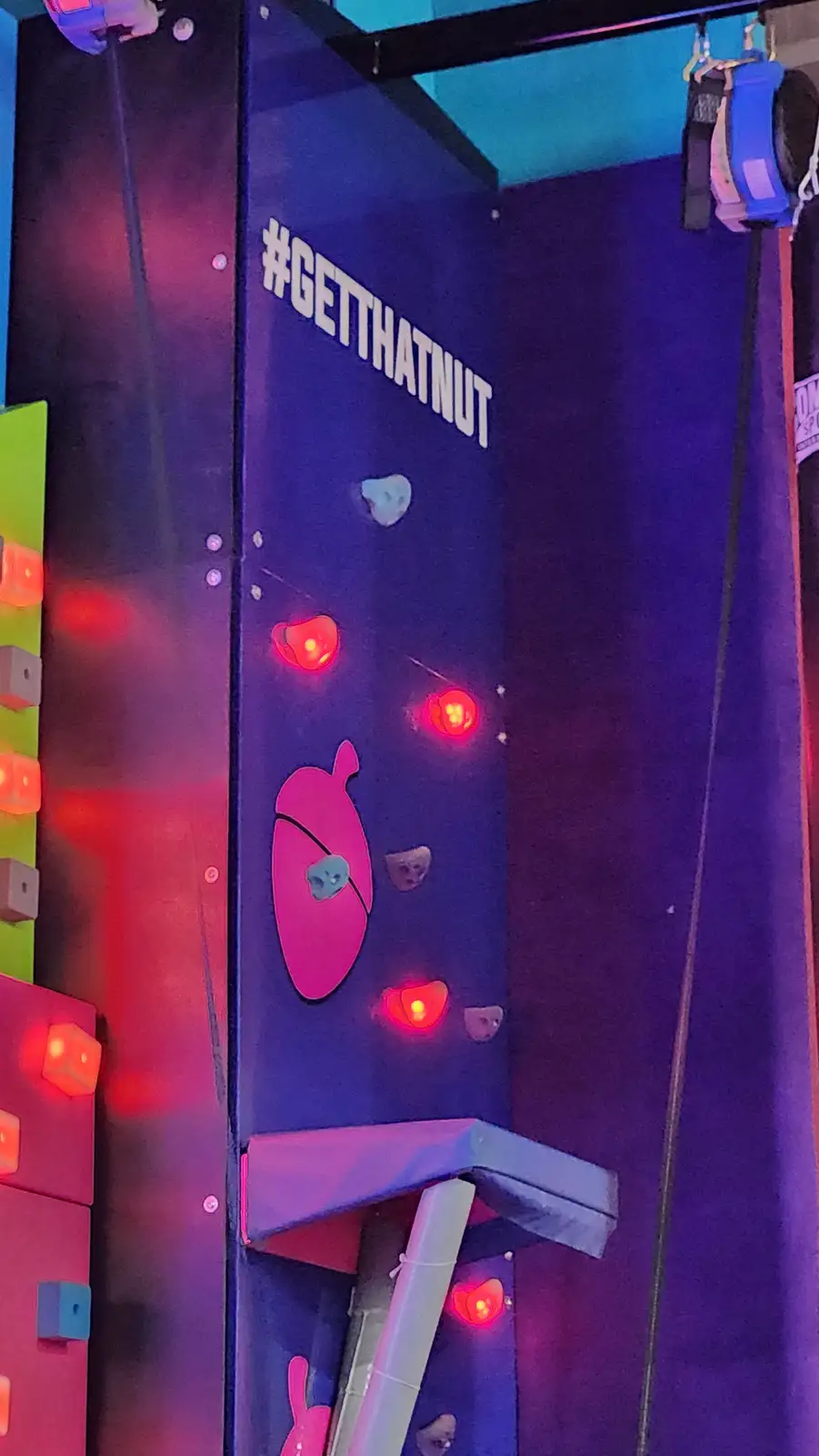 A close-up of an indoor climbing wall with a large pink acorn cutout and multiple glowing red lights. The wall is dark blue…