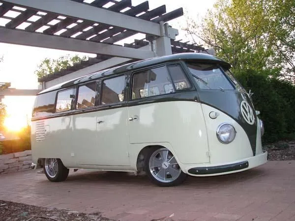 A vintage two-tone Volkswagen Type 2 van, cream and dark green, is parked under a wooden pergola with the sun setting behind…