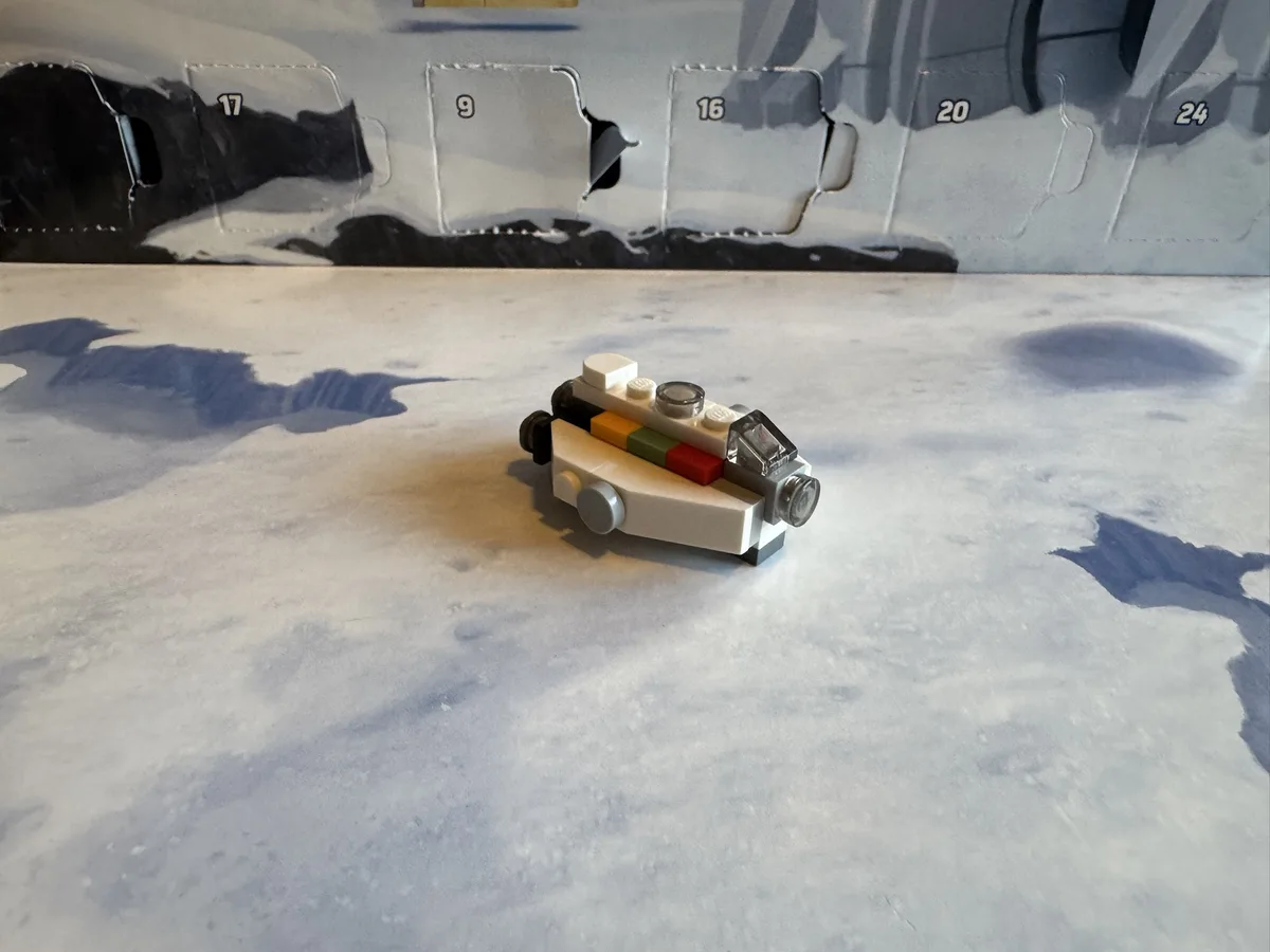 A small, assembled LEGO spaceship model sits on a surface printed with a snowy, mountainous landscape. Behind it, a LEGO…