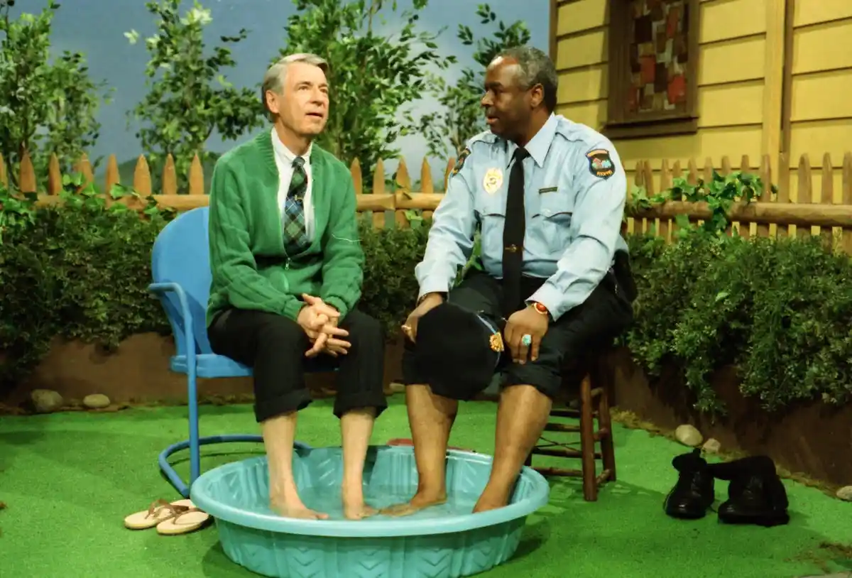 Two men sit on chairs outdoors with their feet in a blue plastic kiddie pool filled with water. The man on the left wears a…