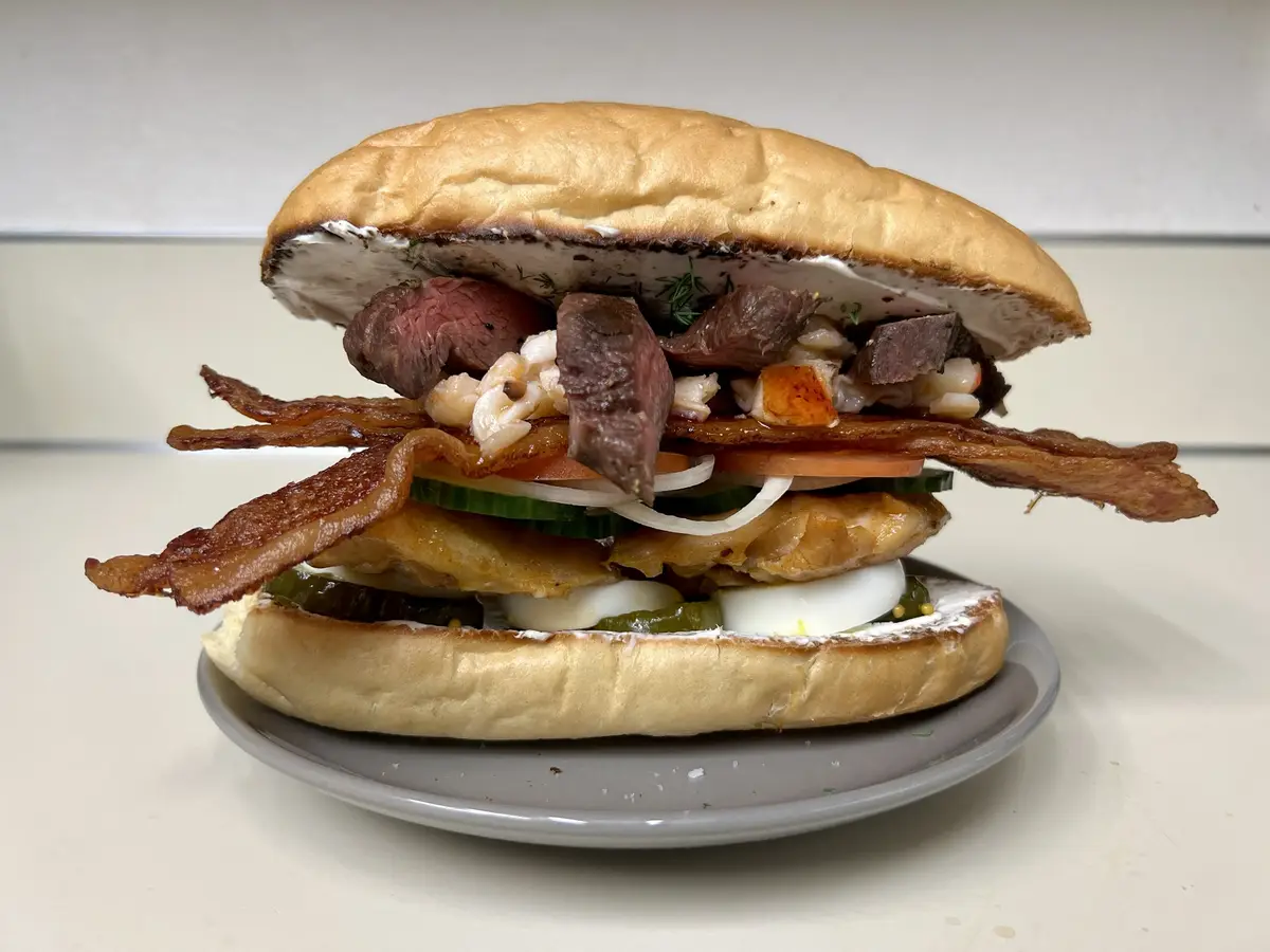 A towering, overflowing sandwich on a bun is piled high with various ingredients including sliced steak, bacon, fried…