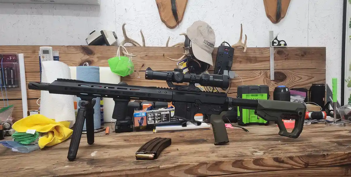 A black rifle with a scope and bipod rests on a cluttered wooden workbench, surrounded by cleaning rags, tools, and a green…