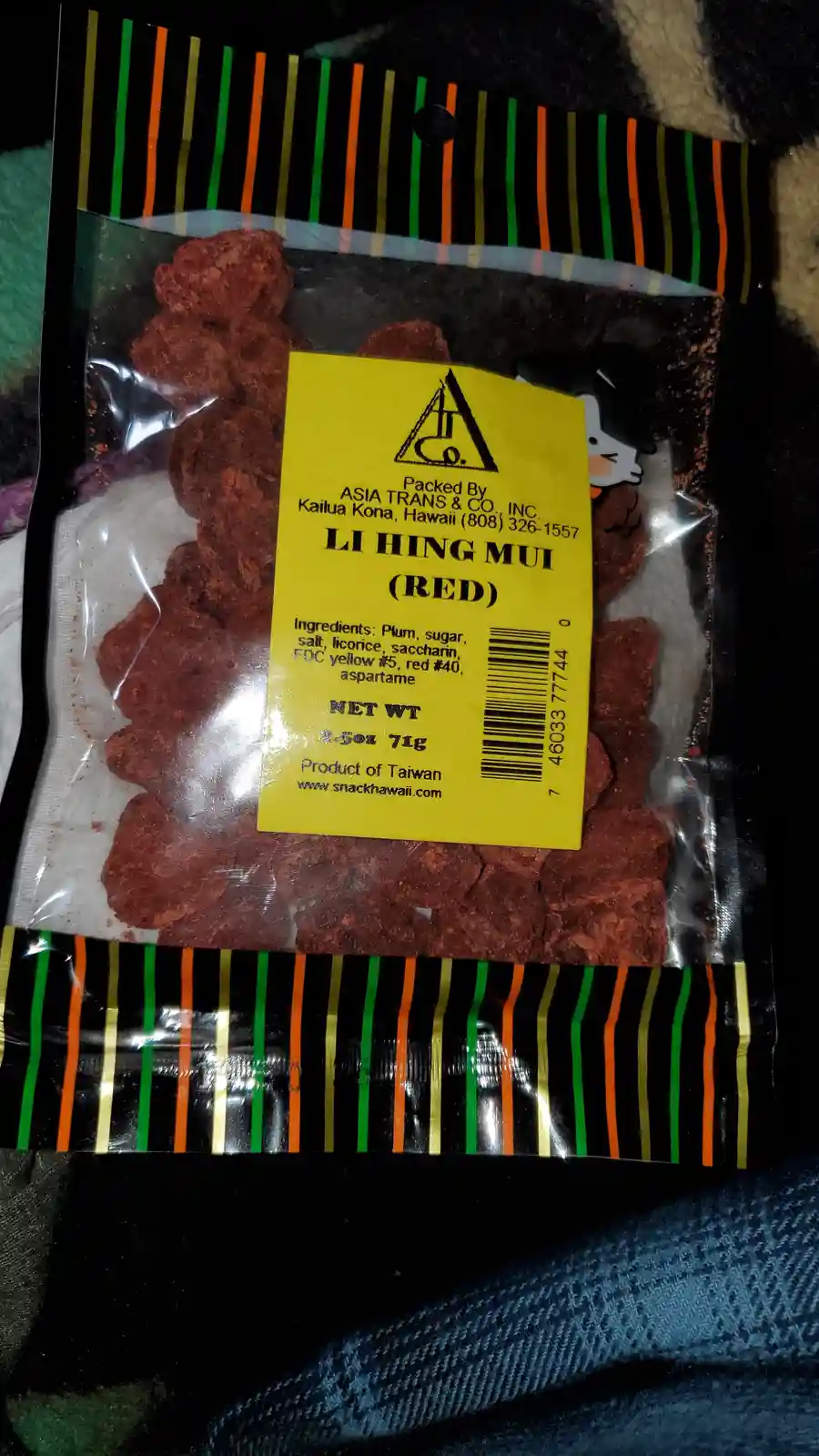 A close-up photo of a plastic bag of red li hing mui snack, showing the yellow label with ingredients like plum, sugar…