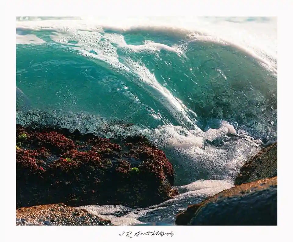 A close-up photograph of a turquoise ocean wave crashing over dark, algae-covered rocks, with white foam at the crest and…