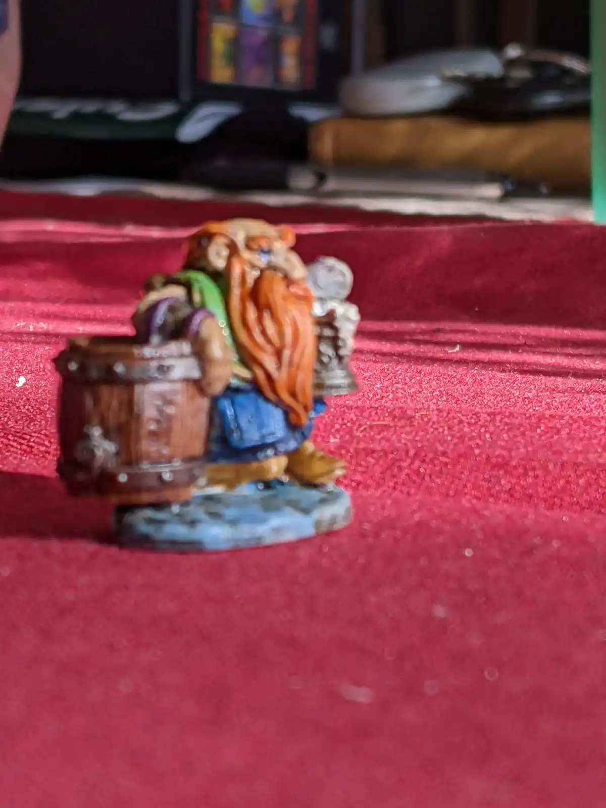 A painted miniature figurine of a dwarf with a long orange beard, wearing blue pants and a green shirt, is shown carrying a…