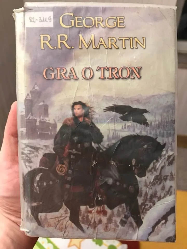 A hand holds a worn, cracked book cover of George R.R. Martin's 'A Song of Ice and Fire' series, with the title incorrectly…