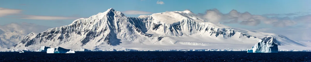 A panoramic view of a snow-covered mountain range under a partly cloudy sky, with several large icebergs floating in dark…