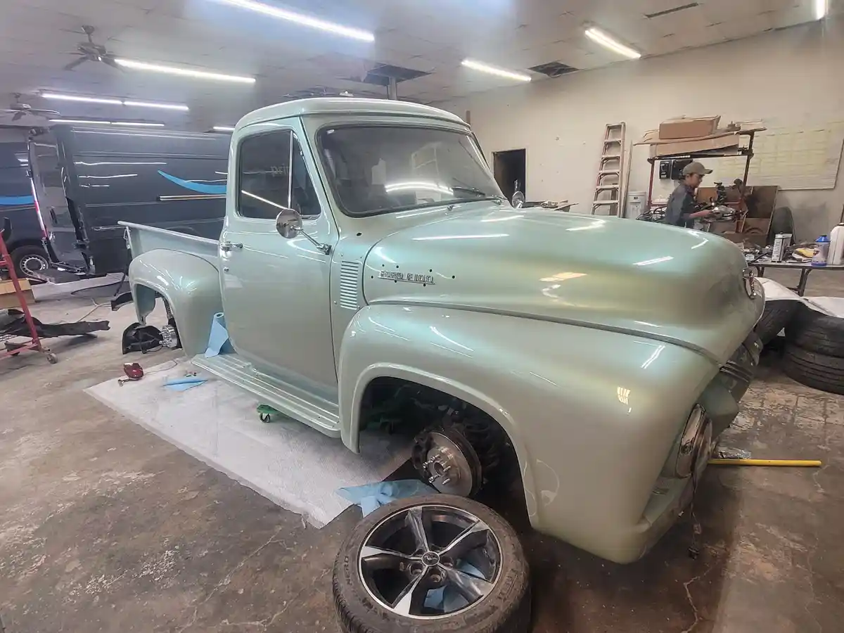 A mint-green vintage Ford F-100 pickup truck is shown in a garage, partially disassembled with its front wheels removed and…