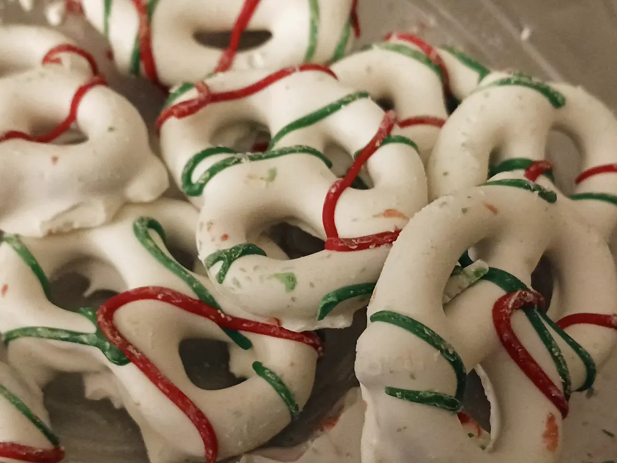 A close-up photo shows numerous white-iced pretzels decorated with red and green candy drizzle, arranged in a pile. The…