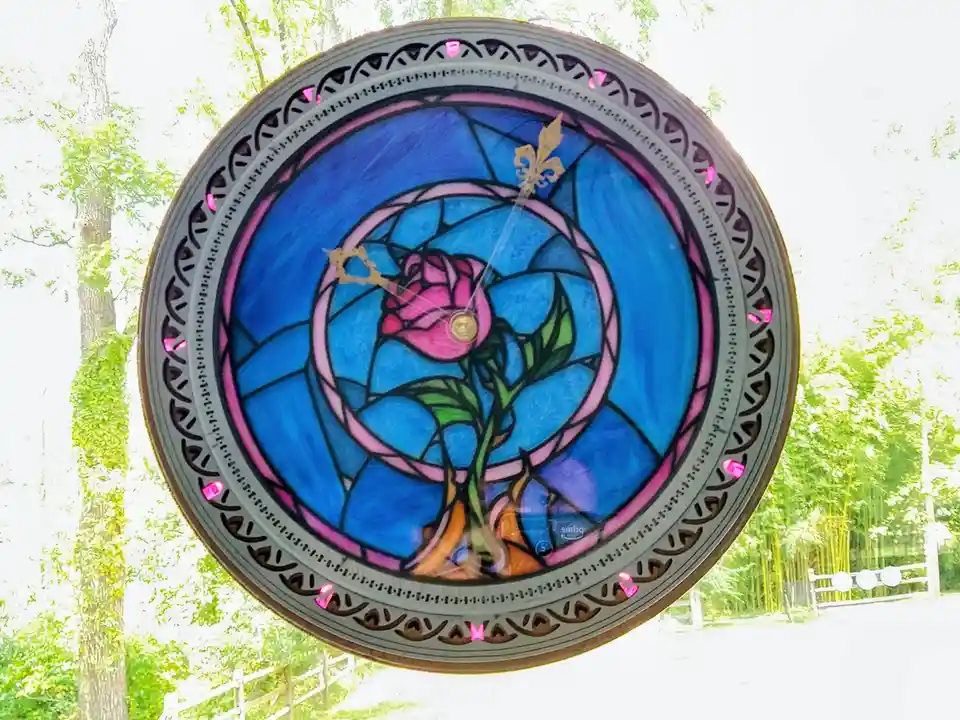 A circular stained-glass clock with a pink rose and blue background, featuring ornate gold hands and pink gem-like hour…