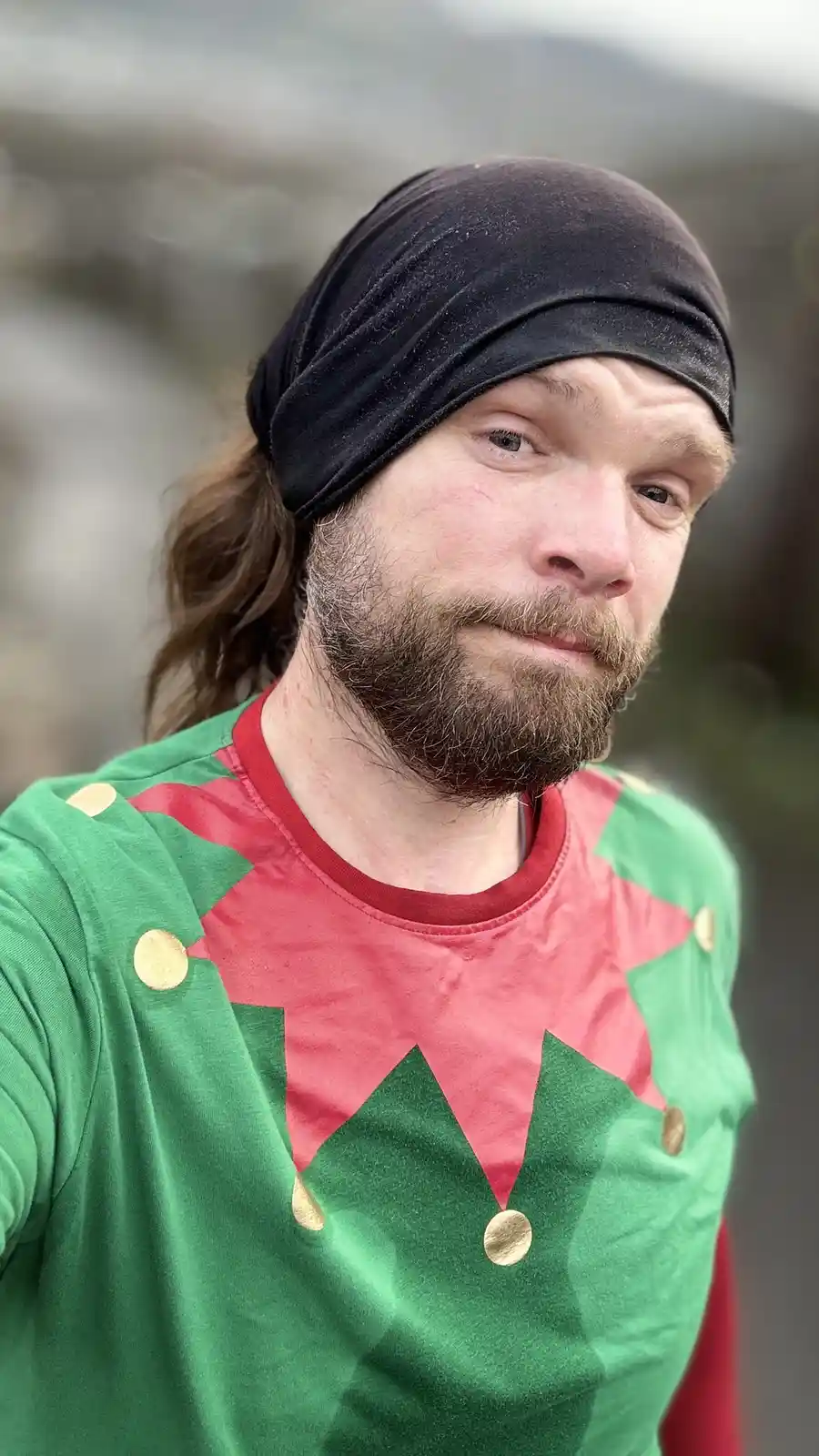 A man with a beard and long hair wearing a black headband and a red-and-green elf costume shirt with gold buttons looks at…
